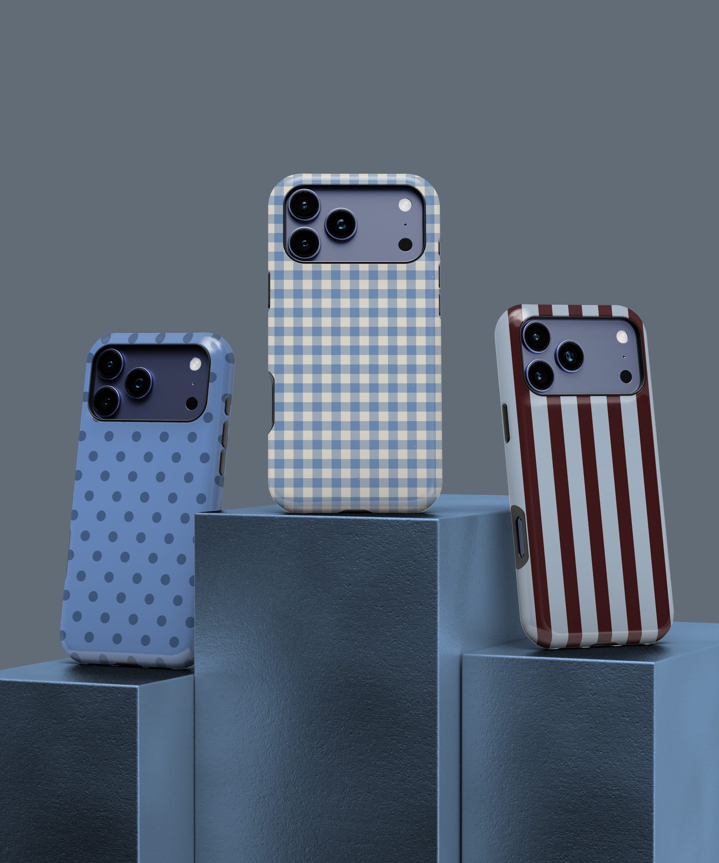 Three phone cases displayed on blue podium blocks: light blue with darker blue polka dots, blue and cream gingham, and cream with vertical red stripes, set against a muted blue background in a minimalist product showcase.